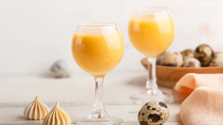 Sweet egg liqueur in glass with quail eggs and meringues on white wooden background. Side view, close up, high key.
