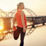 Cute Caucasian blonde woman in sportswear stretching leg by the river. It's hard to beat a person who never gives up.