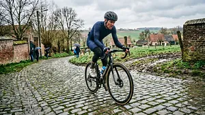 Een actiefoto van een mannelijke wielrenner in de Vlaamse heuvels, die over een steile, natte kasseistrook rijdt. Zijn gezicht toont concentratie en de cobblestones zijn glad van de recente regen. Hij draagt een moderne helm en voorjaarskleding.