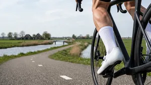 Close-up van het onderbeen van een mannelijke wielrenner in actie, met een perfect zittende witte aero-sok en een moderne witte wielerschoen. De achtergrond toont een onscherp Nederlands polderlandschap