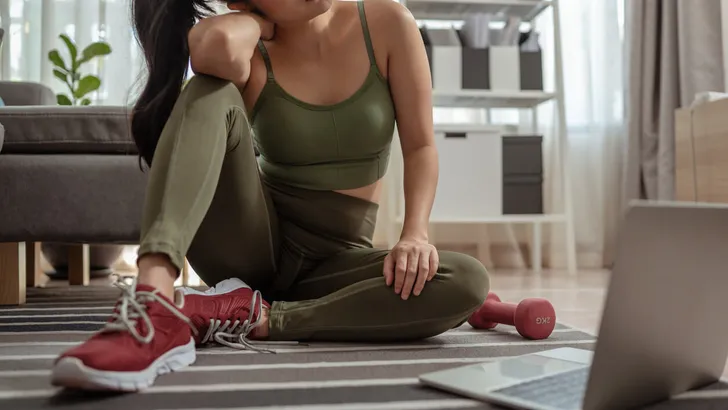 A sporty woman in sportswear is sitting on the floor with dumbbells and is using a laptop at home in the living room.Exercise indoors during quarantine. Exercise, Workout at home activities.