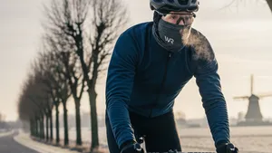 Wielrenner met een zwarte buff en een winterjack fietst op een geasfalteerd fietspad door een typisch Nederlands polderlandschap met knotwilgen en een waterige winterzon.