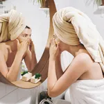 Young happy woman in towel applying organic face scrub and looking at round mirror in stylish bathroom. Girl making facial massage, peeling and cleaning skin on face. Skin Care and Hygiene