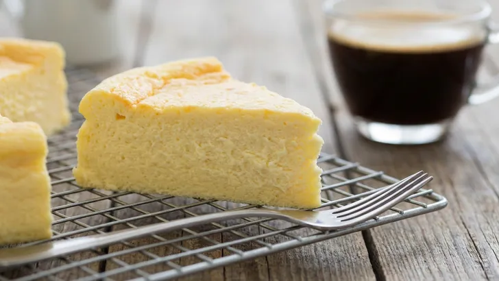 Een close-up van een punt luchtige Skyr cheesecake op een metalen afkoelrek, met een vork ervoor en een kop koffie in de wazige achtergrond op een houten tafel.