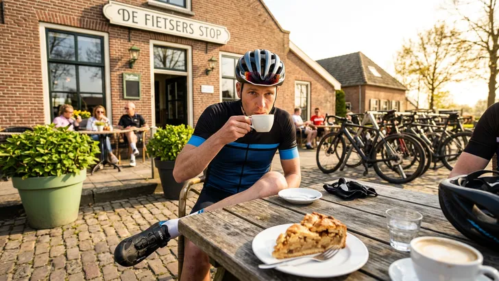 en wielrenner met zijn helm nog op zit op een zonnig terras van een Nederlands fietscafé en kijkt betrapt op van zijn kop koffie, als illustratie van foute terras-etiquette.