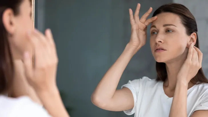 Anxious woman looking in mirror, touching forehead, confused about wrinkles