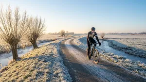 Een eenzame racefietser in volledige winterkleding rijdt over een bevroren, slingerend polderweggetje met rijp op de bomen en velden, onder een heldere winterzon.