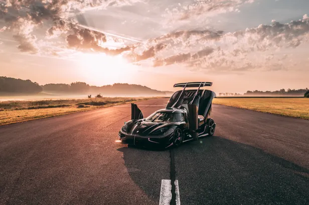 Koenigsegg One:1 met geopende portieren en motorkap op een leeg asfaltterrein bij zonsopkomst.