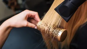 Female client in beauty salon. Close-up of hairdressers hand drying blond hair with hair dryer and round brush, doing new hairstyle