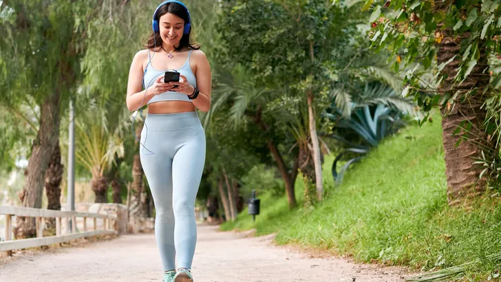 Woman in sportswear walks holding a phone in a city park.