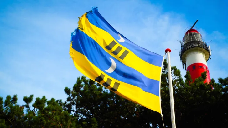 Low angle shot of the Ameland flag flapping in the wind with a lighthouse beside it on a sunny day