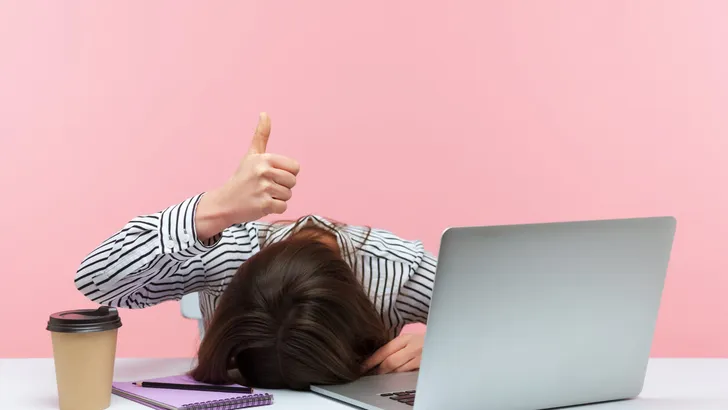 I am fine. Extremely tired sleepy woman lying on table with laptop and cup of coffee showing thumbs up, exhausted with overwork, vitamin deficiency