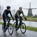 Twee mannelijke wielrenners in winterkleding maken een ontspannen praatje tijdens een rustige duurtraining op een natte weg langs een Hollandse vaart met een molen in de achtergrond.