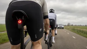 Een mannelijke wielrenner fietst op een open Nederlandse polderweg met een slimme fietsradar inclusief rood achterlicht gemonteerd onder zijn zadel, terwijl een auto hem van achteren nadert.