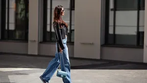 Full length beautiful young brunette woman Wearing black leather jacket and wide blue jeans, sneakers, sunglasses and small handbag with chain, walking street on sunny day