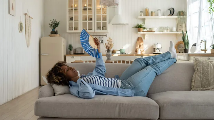 Unhappy exhausted and overheated young African woman lying on sofa at home waving fan to cool down