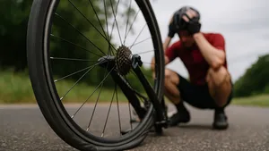Close-up van een racefiets met een volledig lege achterband op het asfalt, met op de achtergrond een radeloze wielrenner die gehurkt langs de weg zit.