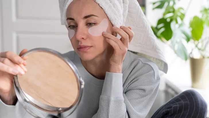 Woman with towel on head apply hydrogel under-eye recovery patches at home, looking in mirror.