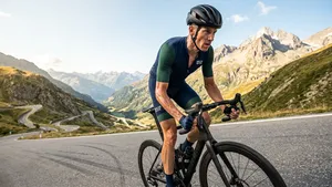 Een afgetrainde mannelijke wielrenner in helm en complete, strakke outfit op zijn racefiets, uit het zadel, op een steile, zonnige bergpas met indrukwekkend Alpenlandschap