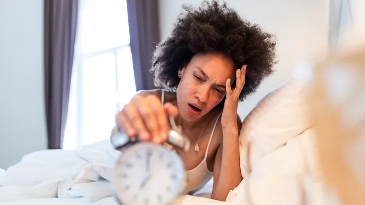 Sleepy young woman stretching hand to ringing alarm to turn it off. Early wake up, not getting enough sleep, getting work concept. waking up with headache