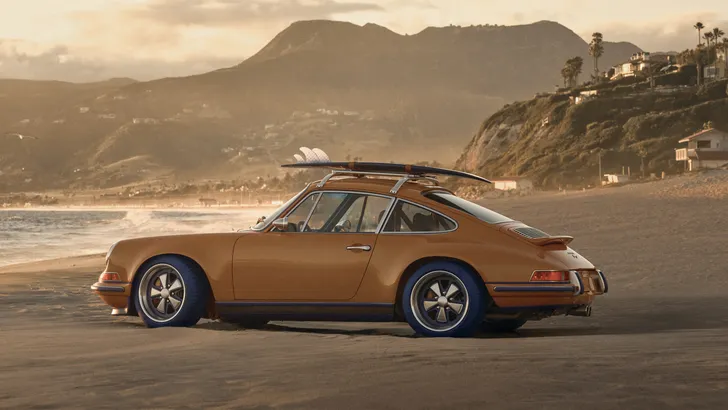 Saffraankleurige Porsche 911 Reimagined by Singer op het strand met een surfplank op het dak.