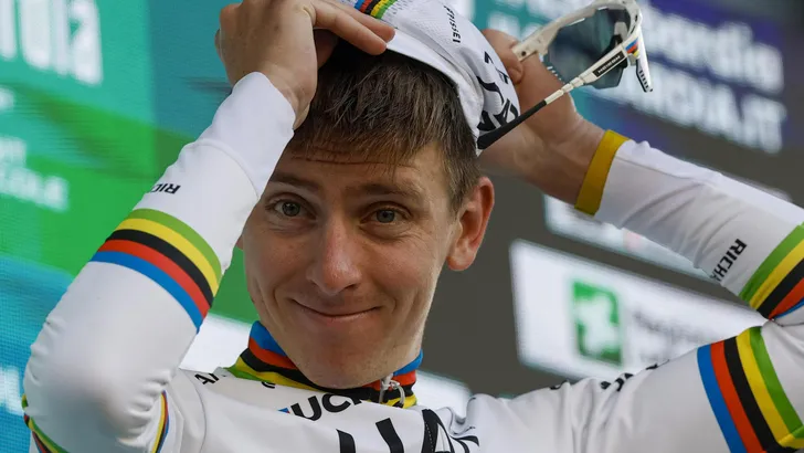 Tadej Pogacar in regenboogtrui glimlacht op het podium en zet zijn pet recht na een overwinning.