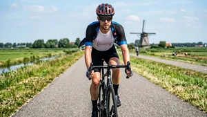 Mannelijke wielrenner met helm op de racefiets in de zon met een opengeritst wielershirt waaronder een wit mesh zweetshirt zichtbaar is.