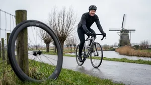 Een lachende wielrenner in winterkleding op een polderweg in Nederland, met een normale racefiets, terwijl een wiel van een duurdere fiets vaag in de voorgrond te zien is.