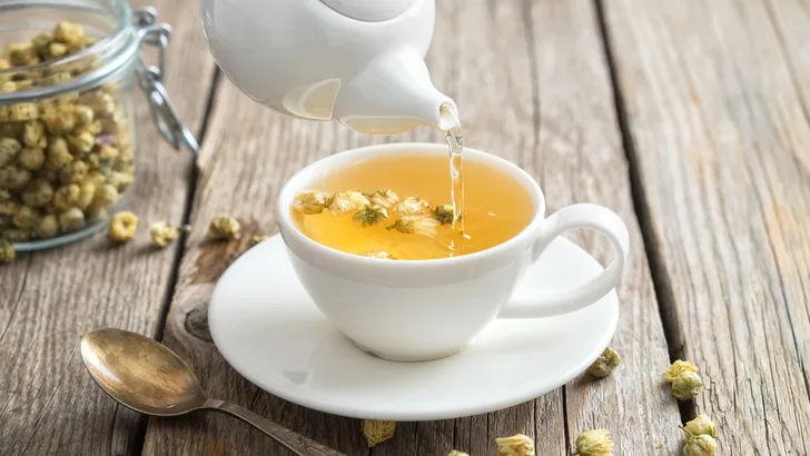 Healthy chamomile tea poured into white cup. Teapot and spoon, glass jar of daisy medicinal herbs.