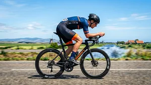 Zijaanzicht van een wielrenner in een diepe, aerodynamische houding, gekleed in een strak fietspak en aero-helm, racend over een open weg.