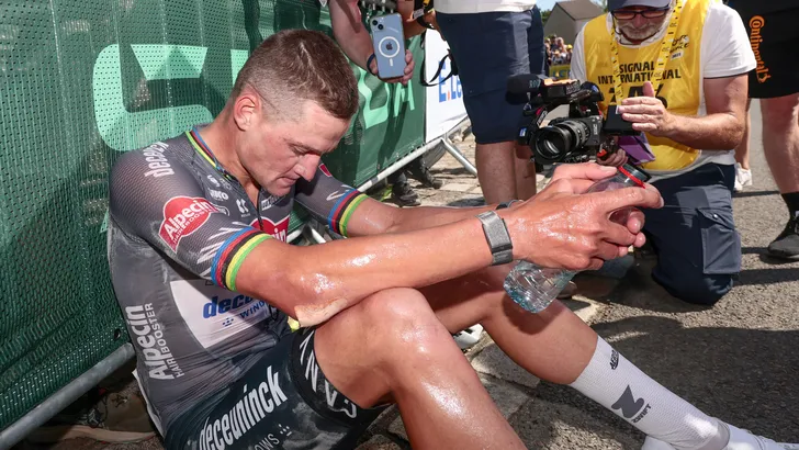 Mathieu van der Poel zit na de finish uitgeput op de grond tegen een dranghek, hevig bezweet en uithijgend na een zware fysieke inspanning in de hitte.
