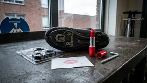 Een set voor het vervangen van schoenplaatjes op een werkbank, bestaande uit een fietsschoen, een open rode lippenstift, een inbussleutel, en nieuwe Shimano SPD-SL cleats.